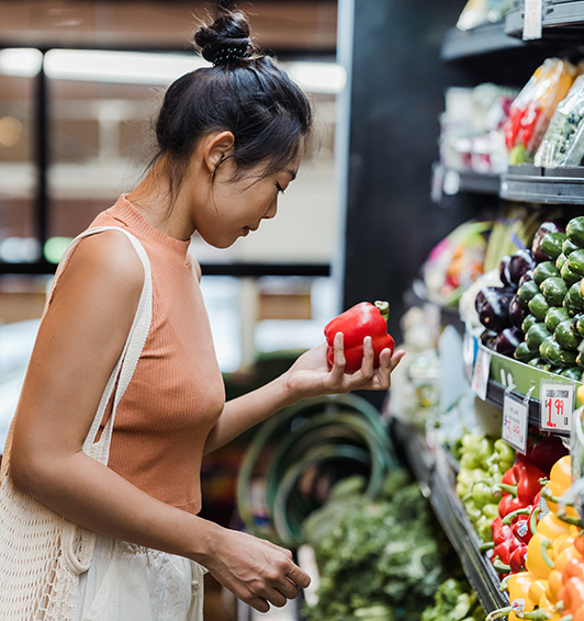 Person with mesh shopping bag holding red pepper in produce section of grocery store.